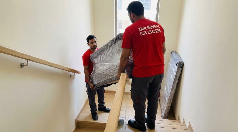 Movers carrying a wrapped bed frame down a townhouse staircase